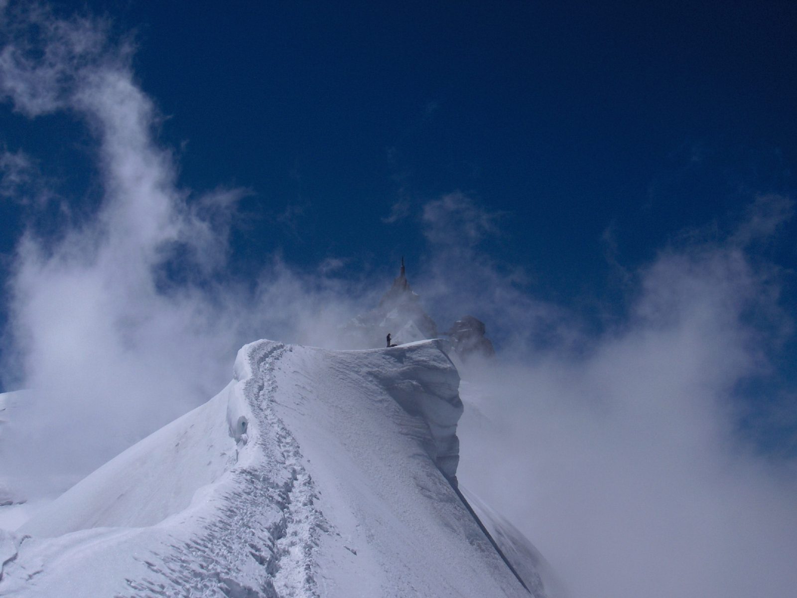 Una arista estrecha de nieve entre nubes, con dos personas diminutas en lo alto