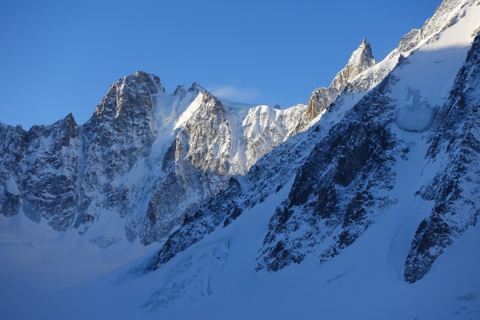 Un glaciar en sombra entre paredes de roca y agujas nevadas, bajo un cielo azul