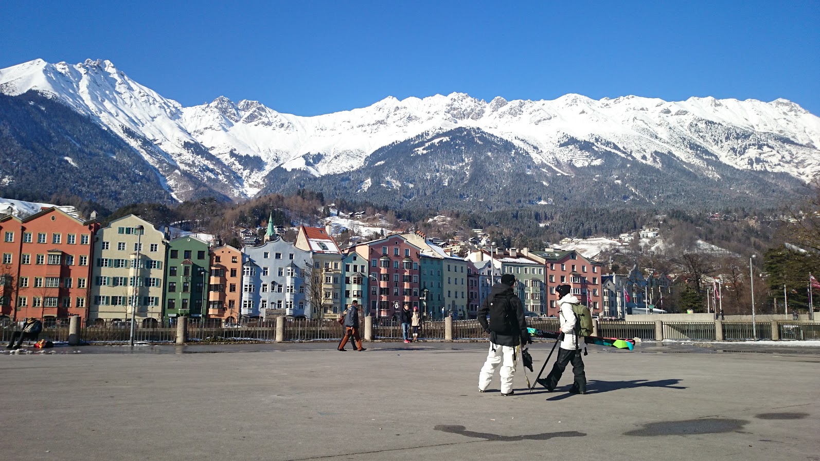 Dos esquiadores caminan con los esquís al hombro por la ciudad, con las montañas nevadas al fondo