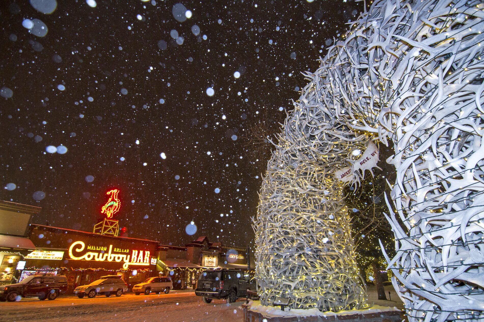 Un arco de astas de wapití iluminado en la plaza de Jackson una noche de nieve, con el letrero luminoso del Million Dollar Cowboy Bar al fondo