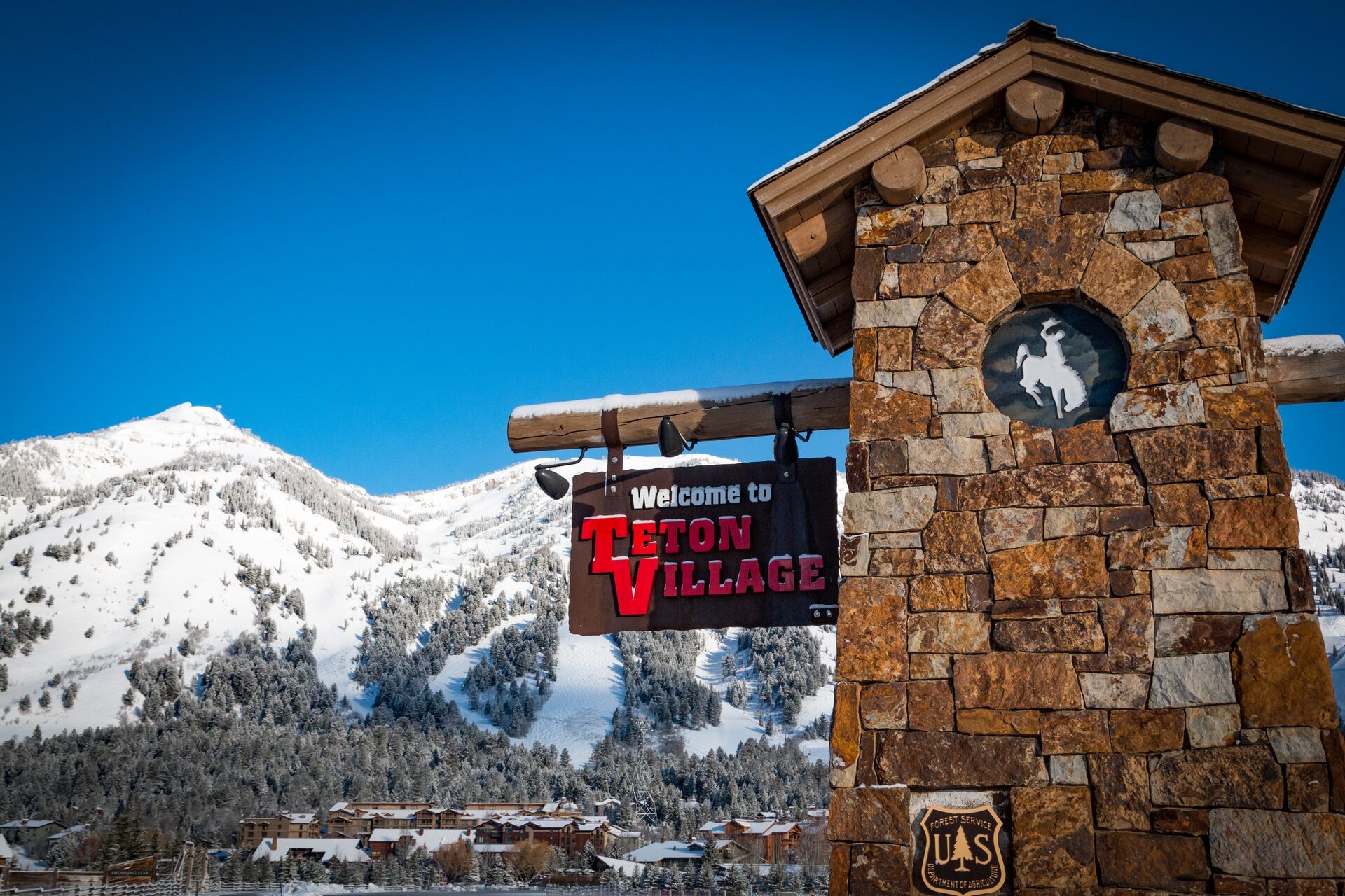 Letrero de bienvenida a Teton Village en un pilar de piedra, con la montaña nevada y los edificios de la base al fondo