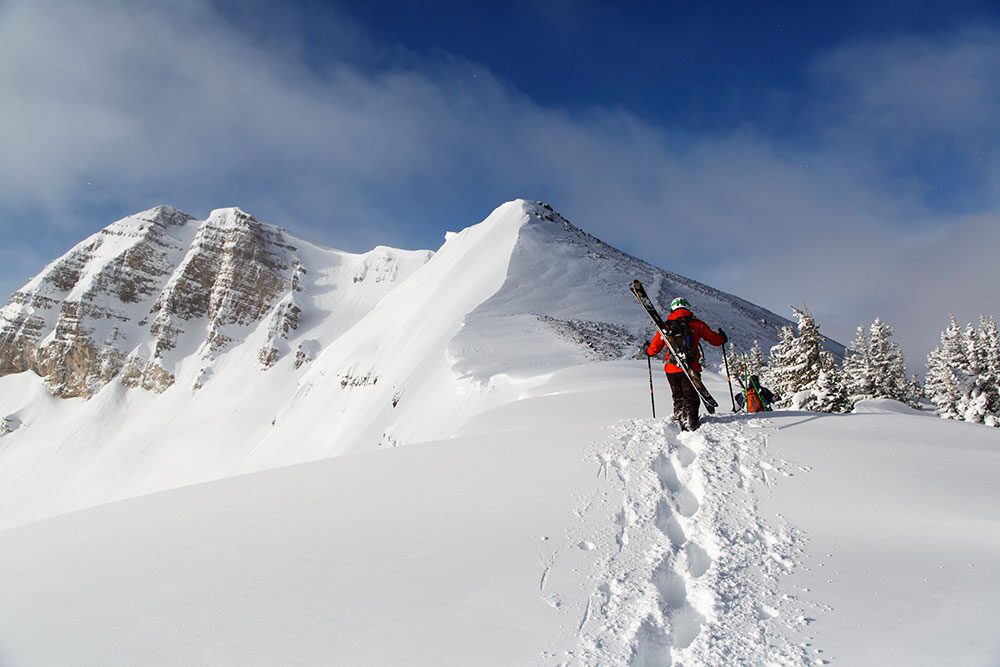 Un esquiador sube a pie por la nieve con los esquís a la espalda hacia una cima nevada y rocosa