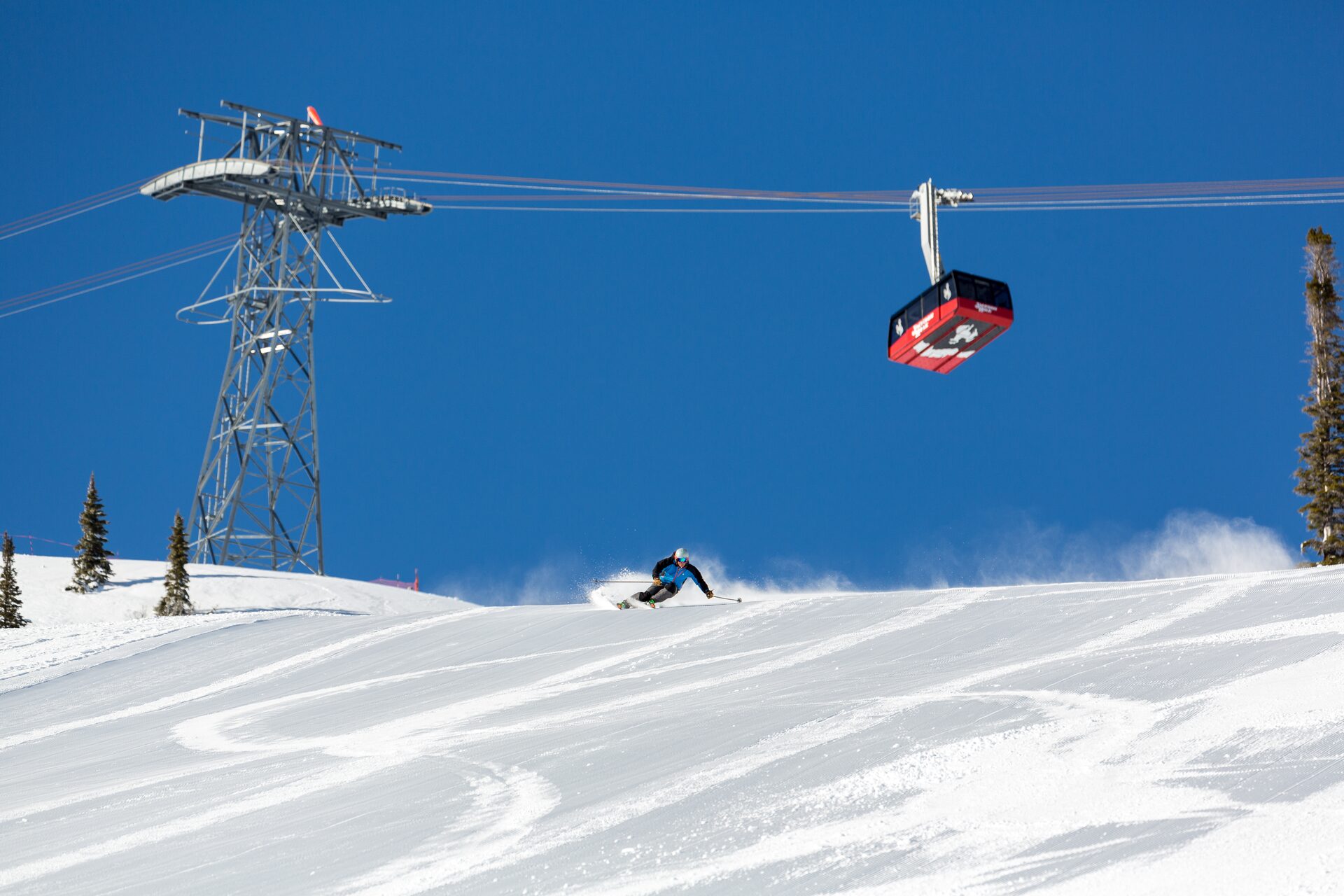 La cabina roja del teleférico de Jackson Hole cruza un cielo azul sobre un esquiador que baja una pista pisada