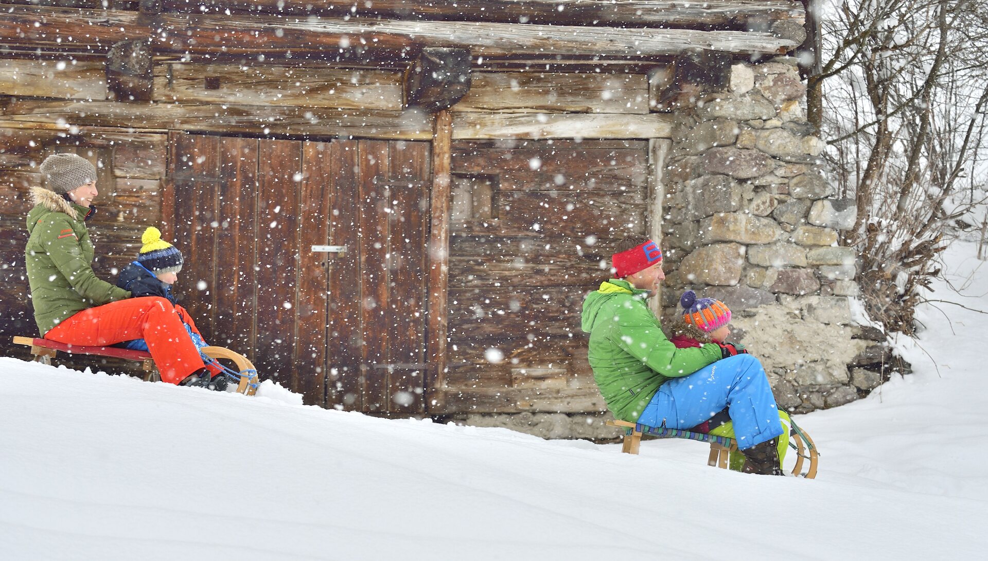 Dos adultos y dos niños bajan en trineos de madera bajo la nevada, delante de un granero antiguo