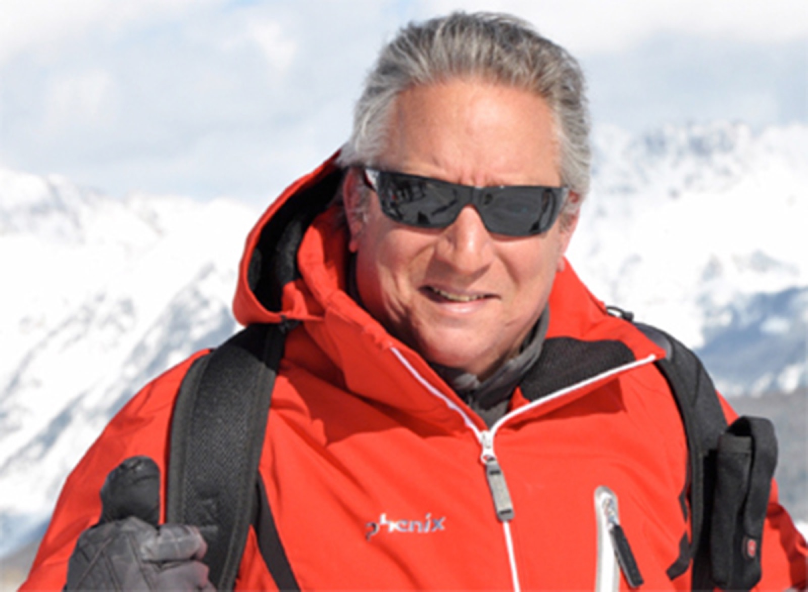 Giora en cumbre alpina, chaqueta roja, gafas de sol, cordillera nevada al fondo